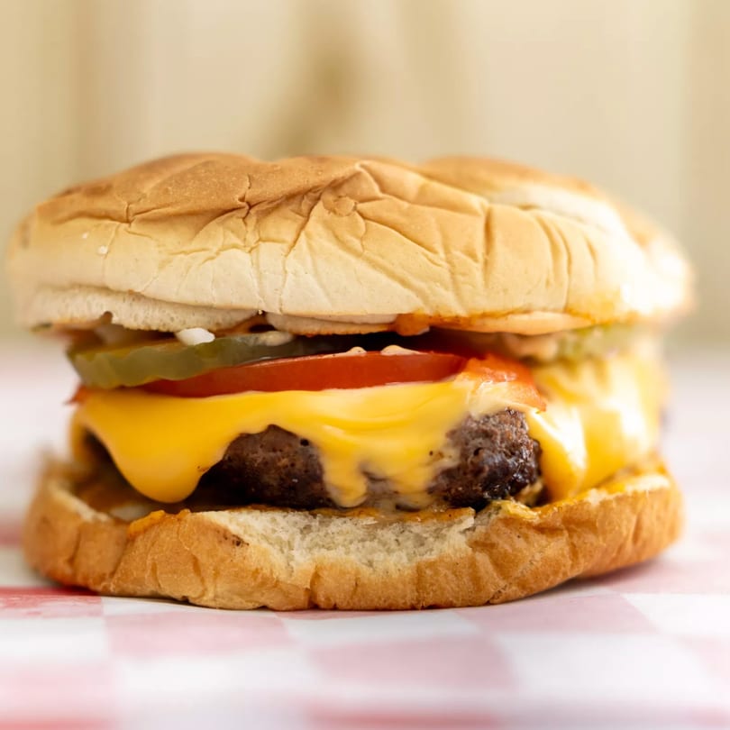 Cheeseburger with melted cheese, tomato, and pickles on a bun, on a red and white checkered paper.