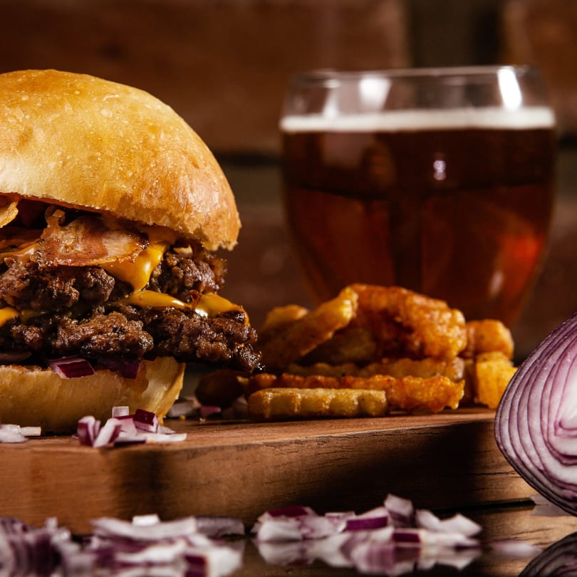 Double patty burger with cheese and bacon, fries, and a beer on a wooden board.